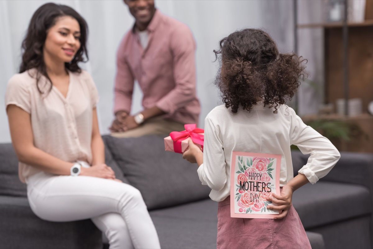 فتاة سوداء صغيرة تقترب من أمها جالسة على أريكة رمادية مع هدية وأم سعيدة