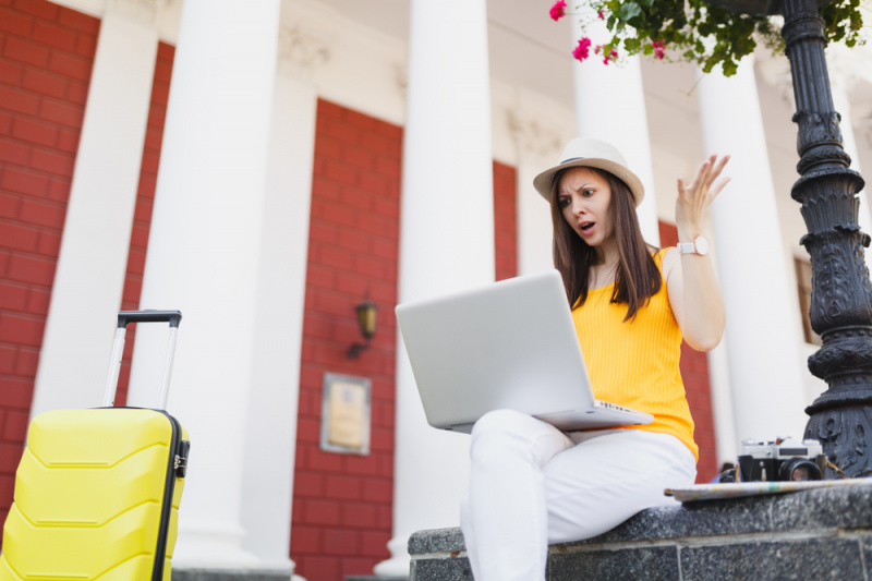   Una joven viajera sentada con su maleta y mirando enojada su computadora portátil