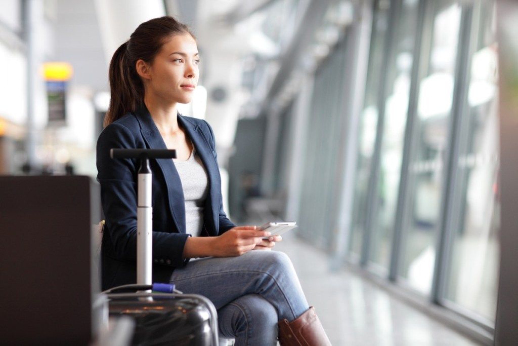 nainen odottaa lentokentällä istumassa matkalaukkujen vieressä älypuhelimen läpi