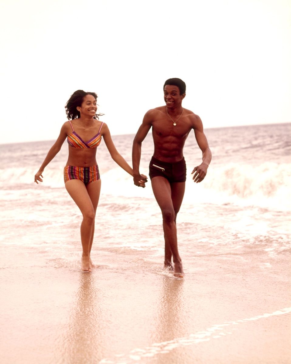 et afroamerikansk par går hånd i hånd på stranden på 1970-tallet