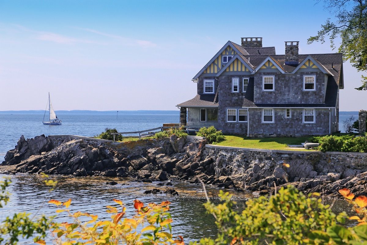 Luksus Waterfront House med grå singel utvendig, Kennebunkport, Maine, New England, USA. Stenete strand, havvann, seilbåt, grønne og gule busker, trær og blå himmel med skyer er i bildet.
