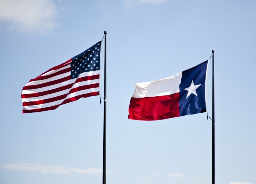Mavi hafif bulutlu bir gökyüzüne karşı birlikte yüksekten uçan Amerika ve Teksas bayrakları