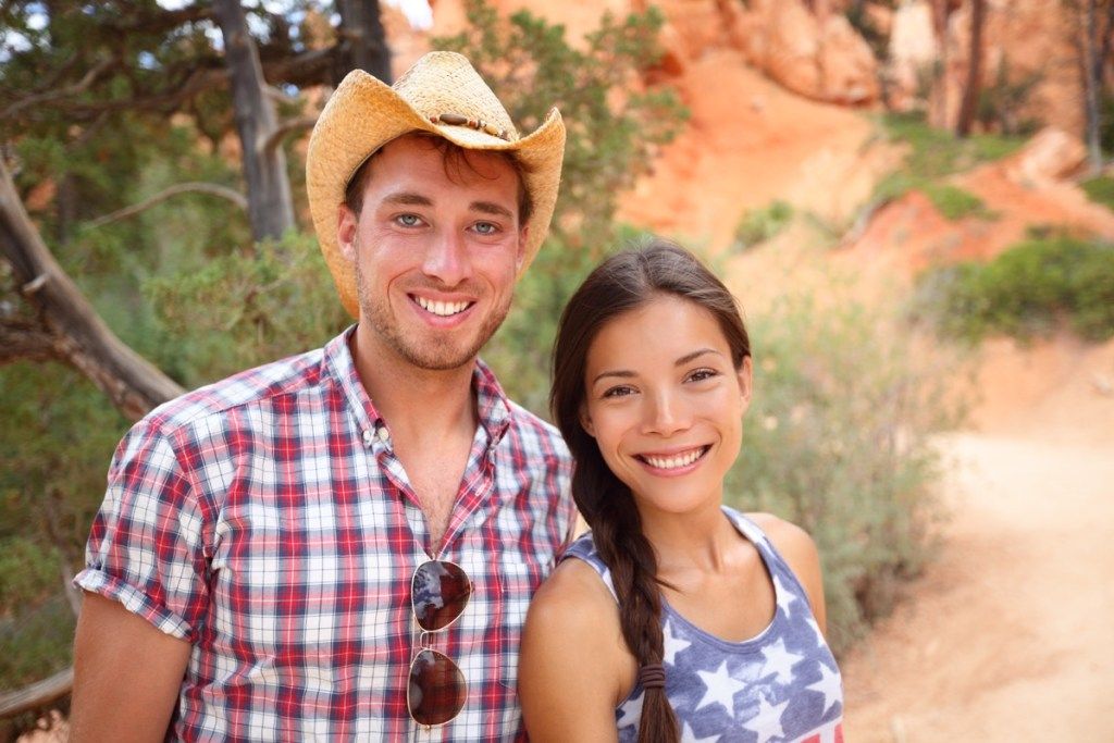 Щастлив портрет на двойка на открито в американска провинция. Усмихната многорасова млада двойка в природата на Западна САЩ. Мъж, облечен в каубойска шапка и жена, облечена в риза на флаг на САЩ