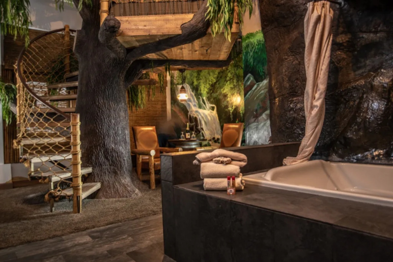   Um quarto de hotel temático com banheira de cachoeira, cadeiras, papel de parede da selva e uma escada para uma cama na casa da árvore